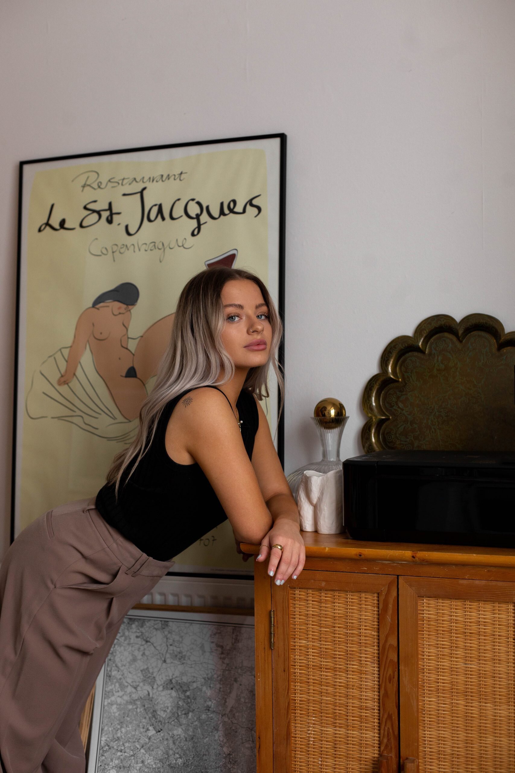 Chloe leans on a wicker cabinet looking into the camera, with an illustration of a nude woman behind her and a vulva vase to the side.